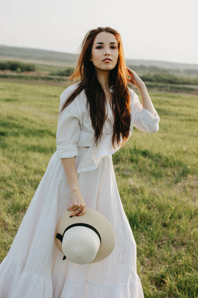 Beautiful carefree long hair asian girl in white clothes and straw hat enjoys life in nature field at sunset. Sensitivity to nature concept - Foto, Imagem