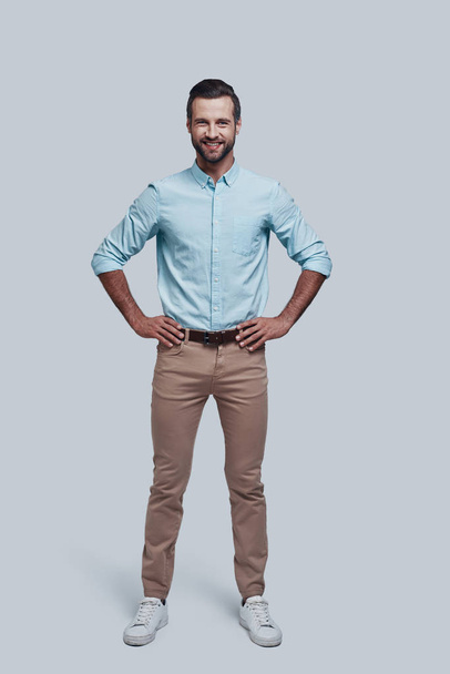 Confidence and charisma. Full length of handsome young man looking at camera and smiling while standing against grey background - Zdjęcie, obraz
