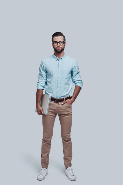 Confident businessman. Full length of good looking young man carrying laptop and looking at camera while standing against grey background - Foto, imagen