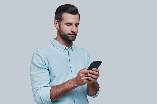 メッセージのチェック灰色の背景に対して立っている間スマートフォンを使用してハンサムな若い男 - 写真・画像