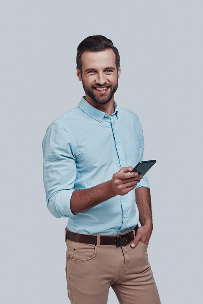 selbstbewusst und erfolgreich. schöner junger Mann mit Smartphone und lächelnd vor grauem Hintergrund - Foto, Bild