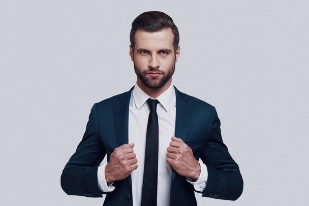 Confident manager. Handsome young man looking at camera and adjusting his jacket while standing against grey background - Foto, Imagen