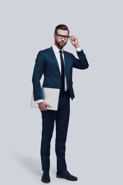 Pewny siebie ekspert biznesowy. Pełna długość przystojnego młodego mężczyzny dostosowując okulary i przewożąc laptopa stojąc na szarym tle - Zdjęcie, obraz