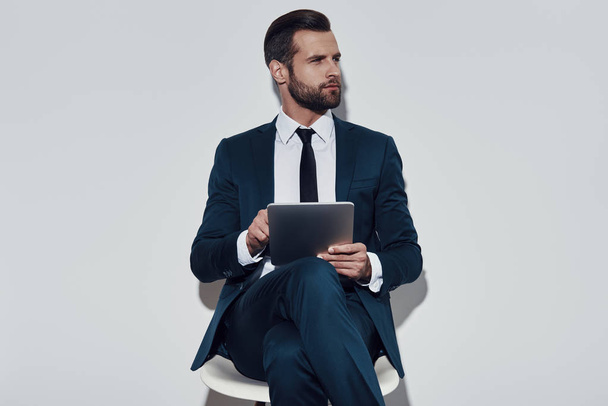 İş yerinde yoğunlaştı. Yakışıklı genç adam uzağa bakıyor ve gri arka plana karşı otururken dijital tablet kullanarak çalışma - Fotoğraf, Görsel