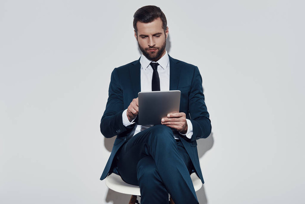 Volle Konzentration. schöner junger Mann arbeitet mit digitalem Tablet, während er vor grauem Hintergrund sitzt - Foto, Bild