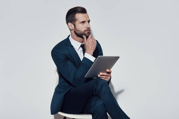 Organizzare tutto. Bello giovane che lavora con tablet digitale e tiene la mano sul mento mentre è seduto sullo sfondo grigio
 - Foto, immagini