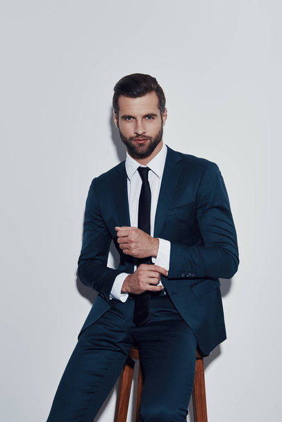 Irresistible man. Handsome young man adjusting his sleeve and looking at camera while sitting against grey background - Foto, Bild