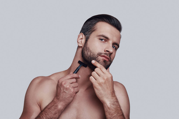 Morning routine. Handsome young man shaving while standing against grey background - Foto, afbeelding