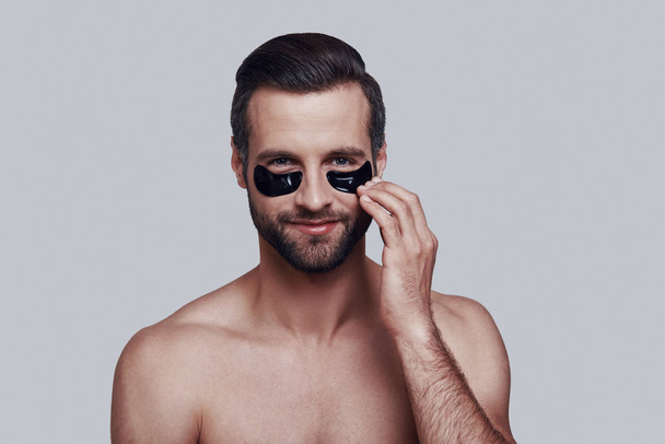 Taking care of his skin. Handsome young man applying medical eye patch and smiling while standing against grey background - Valokuva, kuva