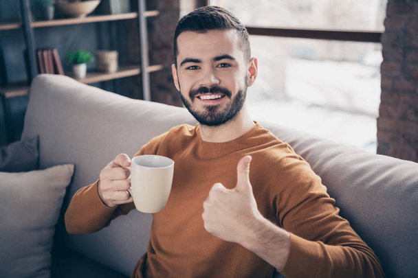 Close-up portrait of his he nice-looking attractive content cheerful cheery bearded guy sitting on divan drinking latte lover showing thumbup recommend at industrial loft interior style living-room - Zdjęcie, obraz
