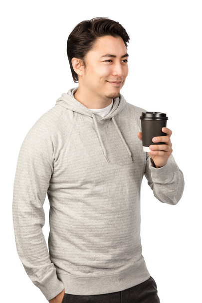 An attractive man in his 20s standing against a white background wearing a grey hood shirt smiling and holding a take-away cup coffee cup, looking away from camera. - Fotografie, Obrázek