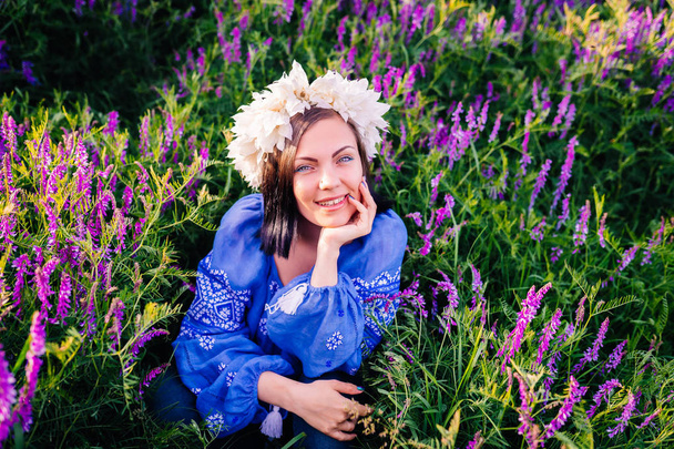 Young pretty woman with flower wreath sitting in purple field. Portrait of girl in blue clothing. - Foto, Imagen