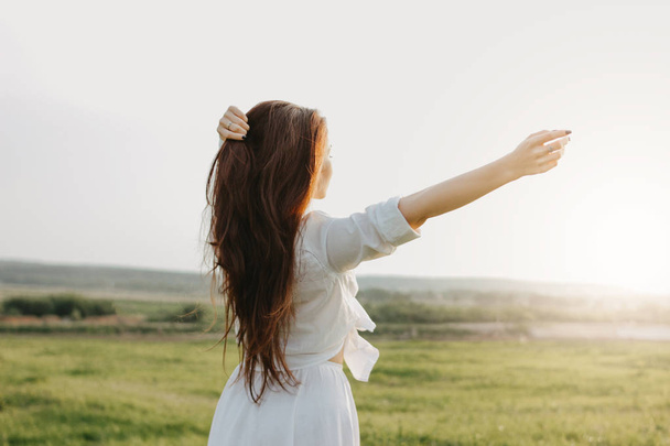 Menina cabelo longo despreocupado em roupas brancas no campo ao pôr-do-sol, vista das costas. Sensibilidade ao conceito de natureza
 - Foto, Imagem