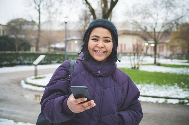 молодая чернокожая женщина стоит в парке со смартфоном и смотрит в окно
 - Фото, изображение