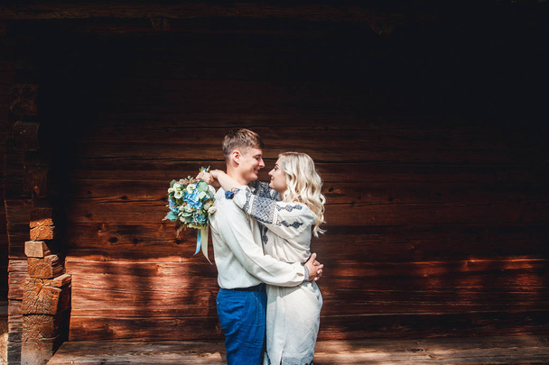 Amazing wedding couple in an embroidereds shirt with a bunch of flowers on the background of a wooden house - Фото, изображение