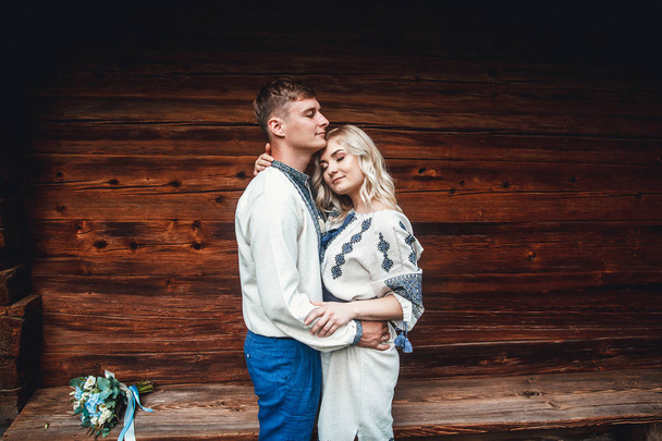 Amazing wedding couple in an embroidereds shirt with a bunch of flowers on the background of a wooden house - Photo, Image