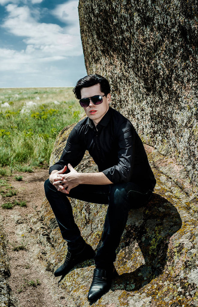 Joven hombre con estilo en traje negro apoyado en enormes rocas
 - Foto, Imagen