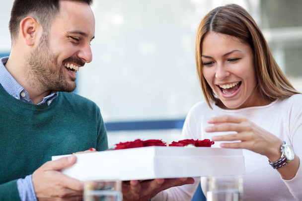 glückliche junge Frau ist überrascht, nachdem sie von ihrem Freund oder Ehemann eine Geschenkbox mit Rosen und Süßigkeiten erhalten hat - Foto, Bild