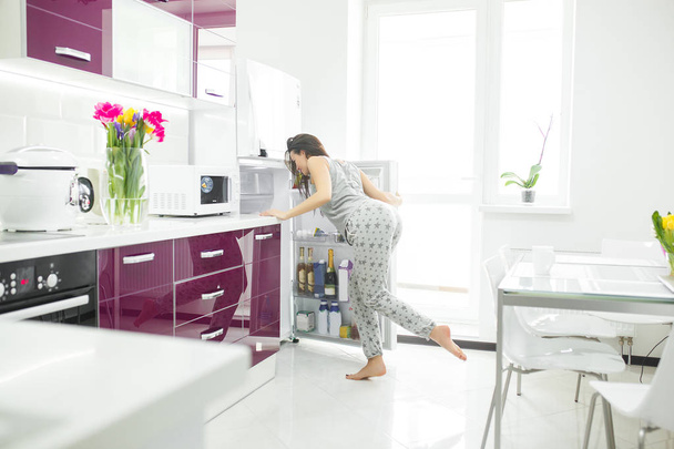 junge Frau in der Küche, die einen Kühlschrank öffnet. Weibchen am Morgen drinnen. Mogelpackung. Vielfraßerin. - Foto, Bild