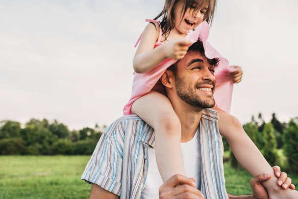 Handsome dad is spending time with his little charming daughter outdoors. Image of happy cute little girl playing with father in the park during the sunset. Fathers Day. Daddy and daughter shares love - Photo, image