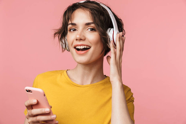 Happy young beautiful woman posing isolated over pink wall background listening music with earphones using mobile phone. - 写真・画像