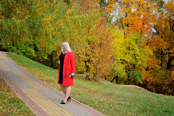 An attractive blonde in a red coat and a scarlet rose in her hand poses on a path in the Park on a Sunny autumn day. Dreaming and looking away. Woman's size plus xxl. Horizontal view - Foto, Imagem