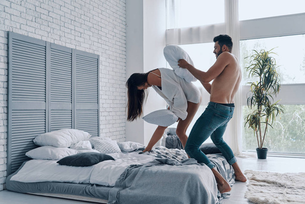 Full length of happy young couple having a fun pillow fight while spending carefree time in the bedroom - Fotografie, Obrázek