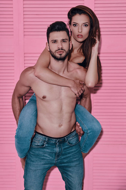 Handsome shirtless man carrying his girlfriend on shoulders while standing against pink background - Foto, imagen