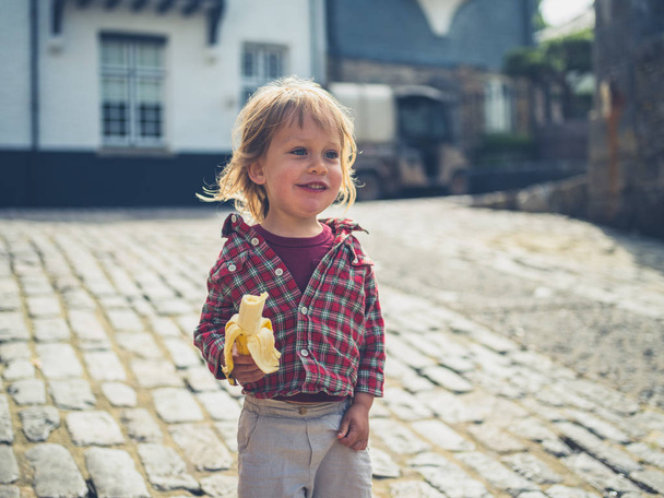 A little toddler is eating a banana outdoors in a cobbled street - Fotó, kép