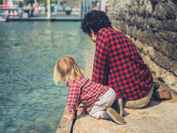 A young father and his toddler are relaxing by the water of a marina - 写真・画像
