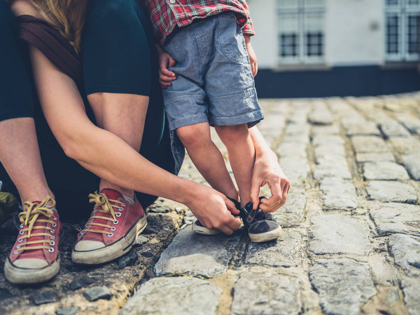 Nuori äiti auttaa pikkulastaan kengät jalassa ulkona mukulakivikadulla.
 - Valokuva, kuva