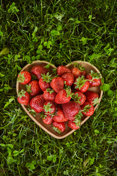 top view of organic strawberries in wooden heart shaped plate on green grass  - Foto, imagen