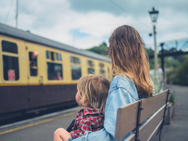 A young mother and her toddler are sitting on a bench at the station watching a train arrive - Valokuva, kuva