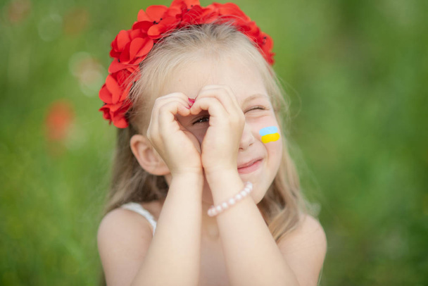 little ukrainian girl looking through heart gesture made with hands in summer green park. Gesture of love to Ukraine by pretty young child in poppy field. - Photo, Image