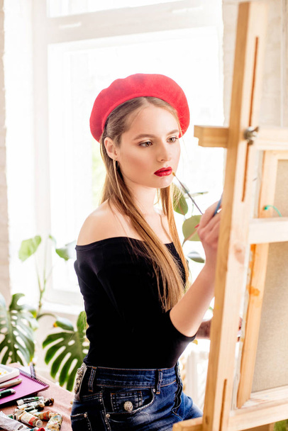 Attractive artist girl with long hair and red beret paints on an easel - Foto, Imagen