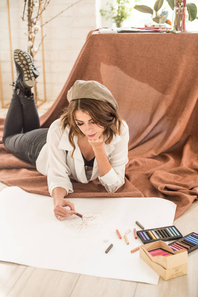 Attractive female artist with dark hair sits on the floor in a studio surrounded by pastel crayons of paper and paints - Foto, Bild