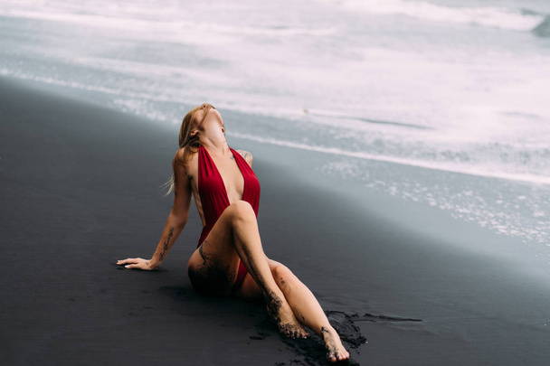 Sexy model with big breasts in a red swimsuit poses on black sand beach. - Photo, image