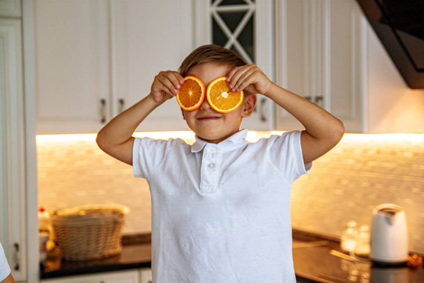 Ολόσωμο πορτρέτο του χαριτωμένο αγοράκι με λευκά ρούχα με πορτοκαλί στέκεται στην κουζίνα. Ιδέα για την υγεία των παιδιών. - Φωτογραφία, εικόνα