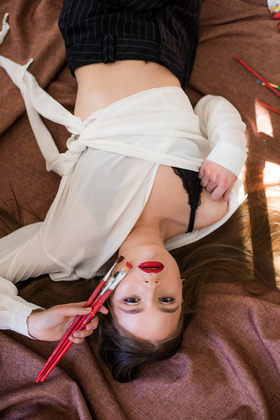 Attractive girl artist with long hair with brushes in hand lies on the floor in the studio - Foto, Imagem