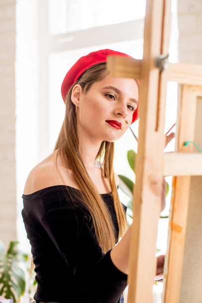 Attractive artist girl with long hair and red beret paints on an easel - Fotoğraf, Görsel