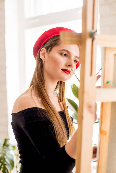 Attractive artist girl with long hair and red beret paints on an easel - Фото, зображення