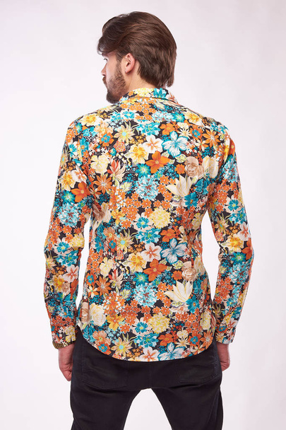 Fashion portrait of young male model with dark hair and beard, posing in floral shirt and black pants. White background - Photo, image