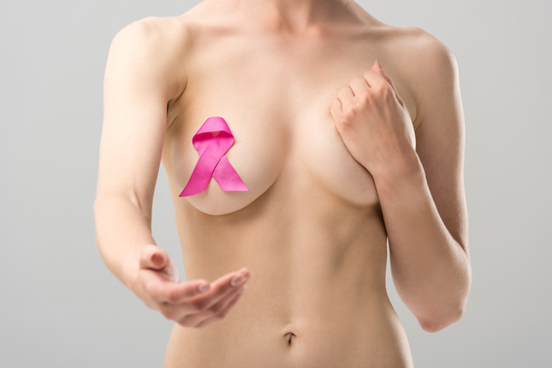 cropped view of naked young woman with pink ribbon with outstretched hand isolated on grey - 写真・画像