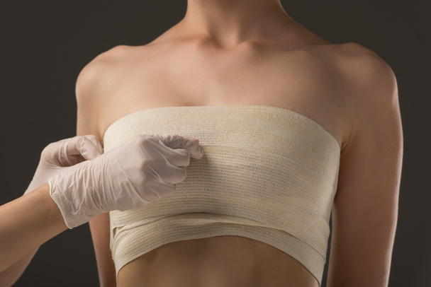 vista parcial del cirujano plástico en guantes de látex y paciente en vendaje mamario aislado en gris
 - Foto, Imagen