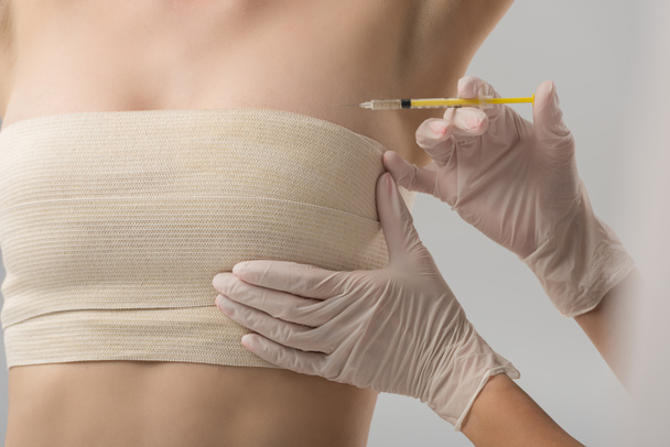 vista parcial del cirujano plástico en guantes de látex inyectables y paciente en vendaje de mama aislado en gris
 - Foto, Imagen