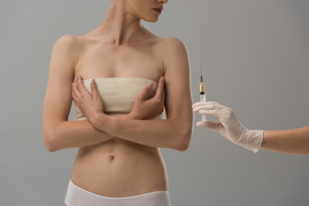 vista recortada de la paciente con vendaje de mama y médico en guante de látex con jeringa aislada en gris
 - Foto, Imagen