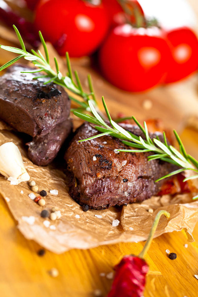Vista ravvicinata di gustose bistecche di cervo alla griglia con rosmarino, aglio, pomodori e spezie in tavola
  - Foto, immagini
