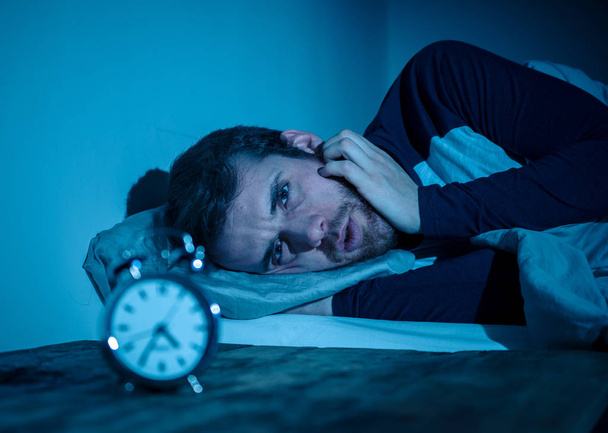 眠れない絶望的な若い白人男性は眠ることができない夜に目を覚まし、ストレスと睡眠障害の概念で不眠症に苦しむ時計を見てイライラし、心配を感じる. - 写真・画像