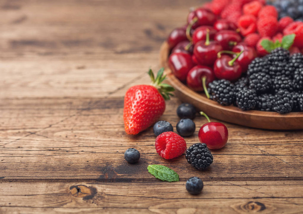 Fresh organic summer berries mix in round wooden tray on light wooden table background. Raspberries, strawberries, blueberries, blackberries and cherries.  - Photo, Image