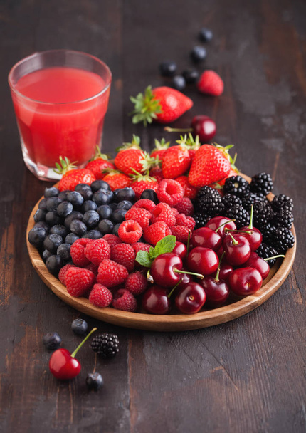 Fresh organic summer berries mix in round wooden tray with glass of juice on dark wooden table background. Raspberries, strawberries, blueberries, blackberries. - Fotografie, Obrázek
